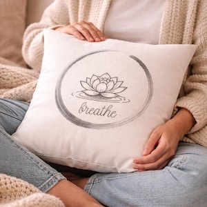 Könnte beinhalten: Ein weißes quadratisches Kissen mit einem grauen Lotusblüten-Design in einem Kreis und dem Wort "breathe" darunter. Das Kissen wird von einer Person gehalten, die ein weißes Hemd, eine helle Strickjacke und blaue Jeans trägt.