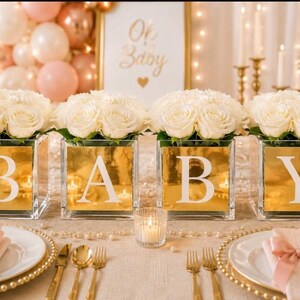 May include: Four square glass vases with a reflective gold finish, each displaying a large white letter to spell BABY. Each vase is topped with a dense arrangement of white roses. The vases are arranged in a row as a centerpiece on a table.