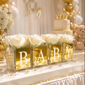 May include: Four square glass vases with gold mirrored fronts, each displaying a large white rose. The vases feature white letters spelling BABY. The arrangement is set on a white table with scattered clear crystals and gold accents.