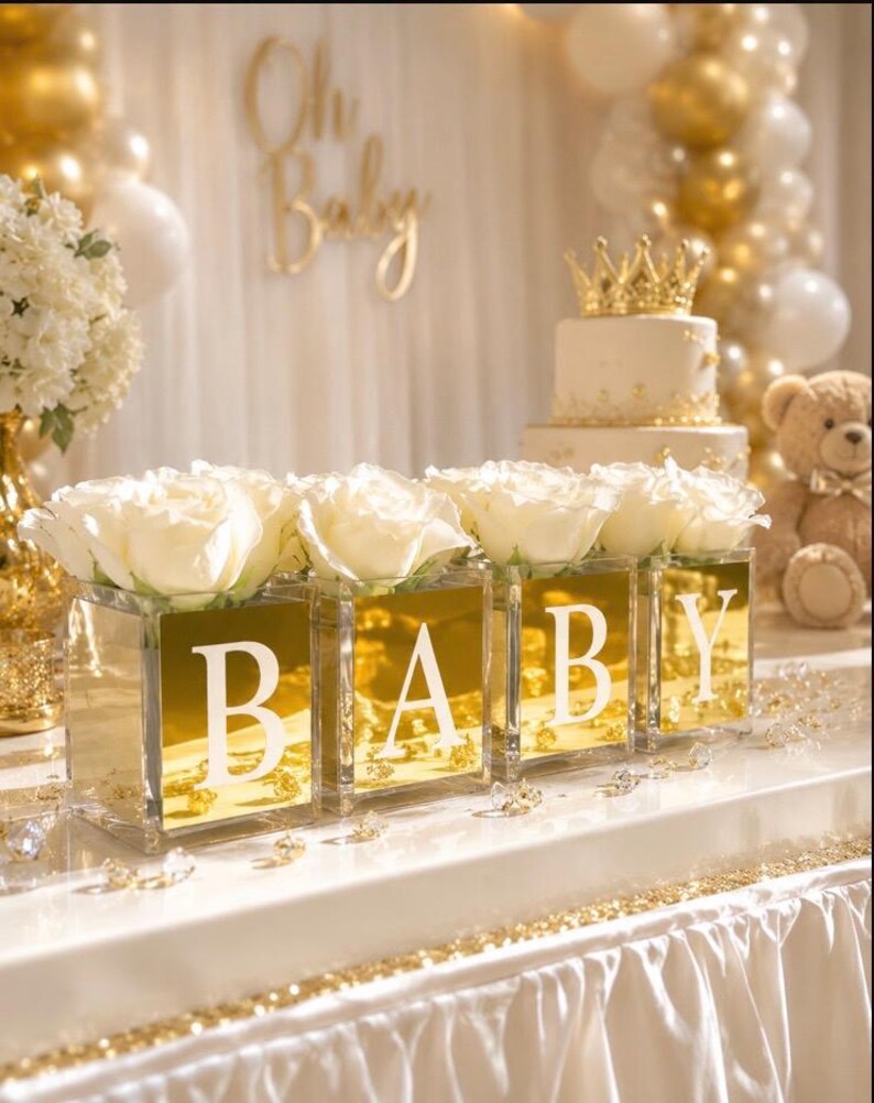 May include: Four square glass vases with gold mirrored fronts, each displaying a large white rose. The vases feature white letters spelling BABY. The arrangement is set on a white table with scattered clear crystals and gold accents.