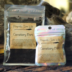 May include: Two clear plastic pouches filled with dark soil, labeled "Magically Clean Witch Cemetery Dirt." The labels feature a witch's broom illustration. Text on the labels describes the product's use in rituals and spells.