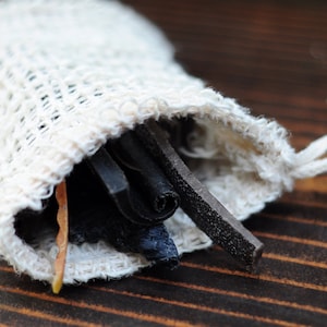 Puede incluir: Una pequeña bolsa de cordón blanco con un borde deshilachado, llena de tiras de cuero negro y marrón. La bolsa está sobre una superficie de madera.