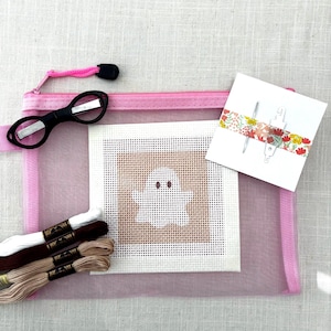 May include: A pink mesh zipper pouch containing a square needlepoint canvas with a white ghost design, a pair of black scissors, three skeins of embroidery floss in white, dark brown, and tan, and a white card with a needle and floral tape.