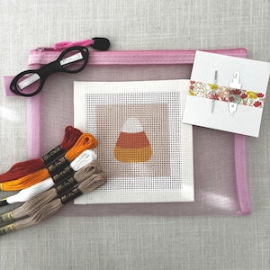 Könnte beinhalten: Ein Stickset bestehend aus einem quadratischen Stramin mit einem Candy-Corn-Motiv, fünf Stickgarnsträngen in Rot, Orange, Weiß und Beige, einer silbernen Nadel, einem Nadeleinfädler auf einer Blumenkarte und einer schwarzen Schere in einer rosa Netztasche.