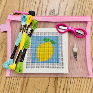May include: A pink mesh zipper pouch containing embroidery supplies. Includes a needlepoint canvas with a lemon design, colorful embroidery floss, pink scissors, and a needle threader. The pouch is on a wooden surface.