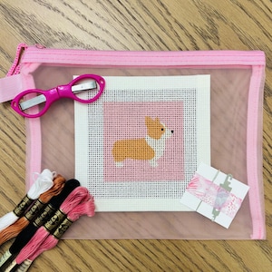 May include: A pink mesh zipper pouch containing a square needlepoint canvas featuring a tan and white corgi on a pink background, a pair of bright pink embroidery scissors, four skeins of embroidery floss, and a needle on a card.