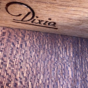 May include: Close-up of a wooden surface with the engraved word "Dixie" in a cursive font. The wood grain is visible, with a darker, textured pattern below the engraved text. The wood appears to be stained a warm brown color.