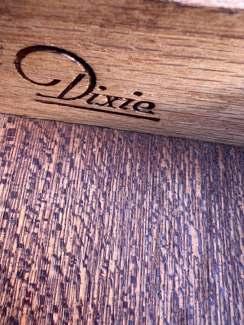 May include: Close-up of a wooden surface with the engraved word "Dixie" in a cursive font. The wood grain is visible, with a darker, textured pattern below the engraved text. The wood appears to be stained a warm brown color.