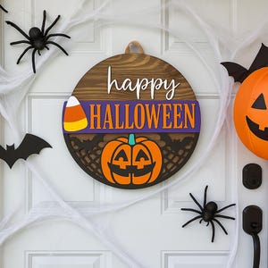 May include: A round wooden Halloween sign with the words "happy HALLOWEEN" in white and orange. The sign features a carved pumpkin and candy corn. Black spiders and bats decorate the white door.
