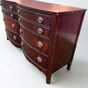 May include: A dark cherry wood dresser with a curved front and six drawers. The dresser has antique bronze hardware and a slightly distressed finish. The top is flat and the legs are short and curved.