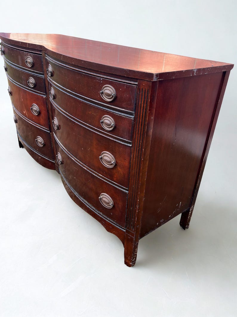 May include: A dark cherry wood dresser with a curved front and six drawers. The dresser has antique bronze hardware and a slightly distressed finish. The top is flat and the legs are short and curved.
