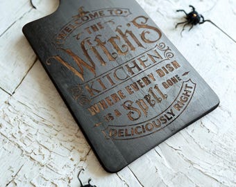 May include: A dark gray wooden cutting board with a handle, featuring engraved text that reads "WELCOME TO THE Witch's KITCHEN WHERE EVERY DISH IS A SPELL DELICIOUSLY RIGHT." Decorative spiders are scattered around the board.