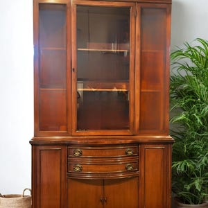 May include: A large, wooden hutch with glass-paned doors and shelves. The lower section features two drawers with brass hardware and two cabinet doors. The wood has a warm, reddish-brown finish, and the piece is set against a white wall.