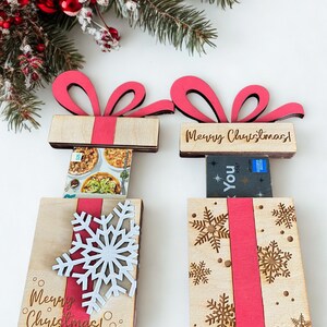 May include: Two wooden gift box-shaped card holders with red ribbon accents and bows. One has a white snowflake, the other has gold snowflakes. Both have the text "Merry Christmas!" and "You". Festive holiday decor.
