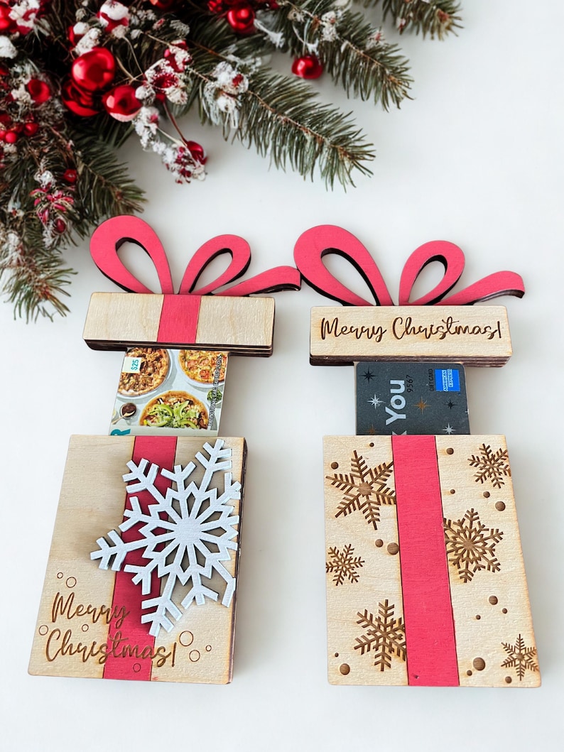 May include: Two wooden gift box-shaped card holders with red ribbon accents and bows. One has a white snowflake, the other has gold snowflakes. Both have the text "Merry Christmas!" and "You". Festive holiday decor.