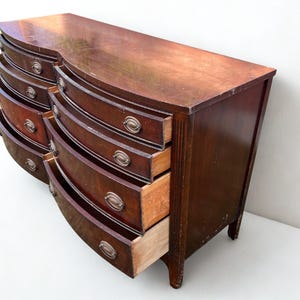 May include: A vintage, dark wood dresser with a curved front and multiple drawers. The drawers have metal hardware and are partially open, revealing the interior. The dresser sits on short legs and has a well-worn appearance.