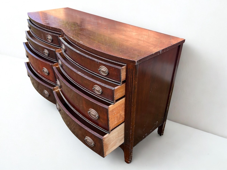 May include: A vintage, dark wood dresser with a curved front and multiple drawers. The drawers have metal hardware and are partially open, revealing the interior. The dresser sits on short legs and has a well-worn appearance.