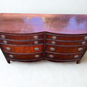 May include: A dark brown wooden dresser with a curved front and six drawers. Each drawer has a silver-colored, oval-shaped handle. The dresser has a slightly curved top and sits on short legs.