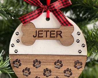 May include: A round, wooden Christmas ornament with a dog bone shape and the name "JETER" engraved on it. The ornament is split into two sections: white and brown, with paw prints and a red and black plaid bow.