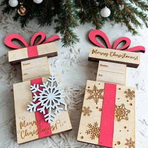 May include: Wooden gift box ornaments with red ribbon and bows. One has a white snowflake, the other has gold snowflakes. Both say "Merry Christmas!" and have space to write "from" and "to".
