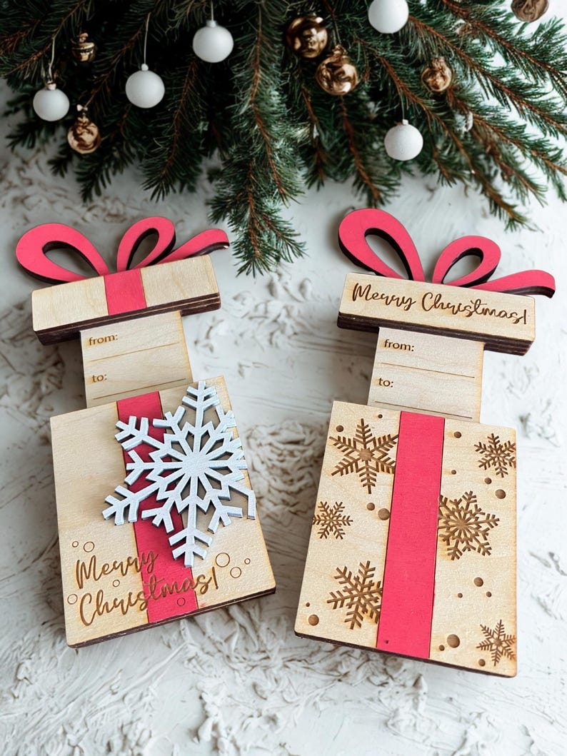 May include: Wooden gift box ornaments with red ribbon and bows. One has a white snowflake, the other has gold snowflakes. Both say "Merry Christmas!" and have space to write "from" and "to".