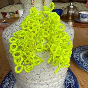 Puede incluir: Pendientes de color amarillo neón brillante con un diseño delicado y de encaje. Los pendientes están suspendidos de ganchos dorados. Se muestran sobre un jarrón blanco y gris moteado, con un plato estampado en azul y blanco en el fondo.