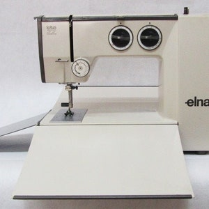 Puede incluir: Una máquina de coser Elna Lotus 822 blanca con una mesa de extensión abatible. La máquina tiene un diseño negro y plateado con dos diales en la parte delantera.