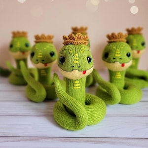Op de afbeelding: Een groep handgemaakte groene gebreide slangen, elk met een kleine bruine kroon. De slangen hebben zwarte ogen, rode tongen en gele accenten. De speelgoedslangen zijn geplaatst op een lichtgekleurd oppervlak.