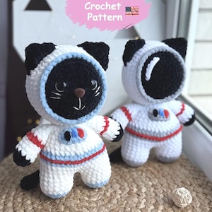 May include: Two crochet cat astronaut dolls. The dolls are white with black accents, wearing astronaut suits with red and blue stripes. The text "Crochet Pattern" is above the dolls.