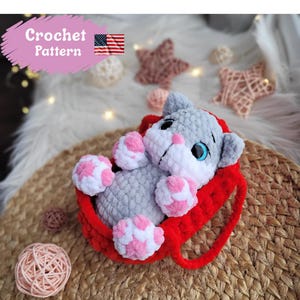 Peut inclure: Un chat en crochet gris avec des pattes roses et blanches et un nez rose repose dans un panier en crochet rouge. Le chat a les yeux bleus. L'image comprend le texte "Crochet Pattern" et un petit drapeau américain.