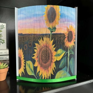 Op de afbeelding: Een boekkunst sculptuur van drie zonnebloemen in een veld bij zonsondergang. De zonnebloemen zijn gemaakt van gevouwen pagina's van een boek en zijn verticaal gerangschikt. Het beeld staat op een witte basis met een groen lint.