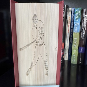 Könnte beinhalten: Buchskulptur mit einem Baseballspieler in Aktion, hergestellt durch Falten der Seiten. Das Buch hat einen roten Buchrücken und Kanten, mit einem hellblauen Band am unteren Rand. Das Kunstwerk wird auf einem Regal mit anderen Büchern ausgestellt.