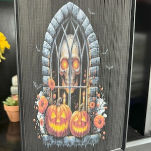 May include: A black book with a skull and pumpkins in a window. The book is folded into a 3D shape.