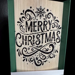 Puede incluir: Una obra de arte hecha a mano con un libro, con las palabras "MERRY CHRISTMAS" cortadas en las páginas. El texto es negro, rodeado de adornos decorativos y un copo de nieve. El libro está en un marco verde con una cinta blanca.
