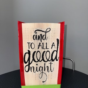 Op de afbeelding: Een handgemaakt boekkunstwerk met de woorden "and to all a good night" uitgesneden in de pagina's. De randen zijn rood en er is een groen lint aan de basis. Het boek wordt op een zwart oppervlak gepresenteerd.