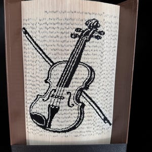 May include: A decorative book sculpture featuring a black and white violin and bow design. The artwork is crafted from folded book pages, set within a brown frame. The violin's details are clearly defined against the textured background of the pages.
