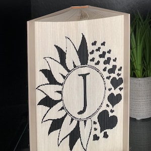 May include: A decorative book with folded pages, featuring a black sunflower design with a central letter "J" inside a circle, surrounded by black hearts. The book has a beige cover and is tied with twine.