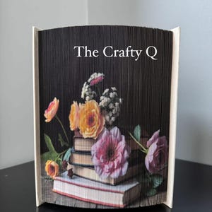 Può includere: Un libro decorativo con un design floreale. Le pagine del libro sono piegate per creare una forma unica, con un'immagine floreale di rose e altri fiori. Il testo "The Crafty Q" è stampato sul fronte.