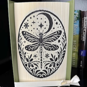 May include: A decorative book art piece featuring a black dragonfly design within an oval frame, set against a backdrop of folded pages. The design includes a crescent moon, stars, and floral elements. The book is held open with a white ribbon.