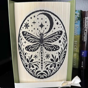 May include: A book sculpture with a black dragonfly design within an oval frame, set against folded pages. The design includes a crescent moon, stars, and floral elements. The book is held open with a white ribbon. The book is displayed on a shelf.