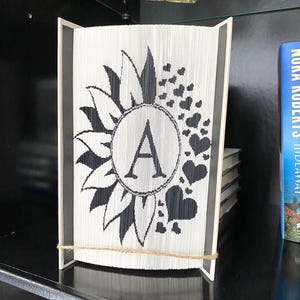 May include: A white book folded into the shape of a sunflower with a black letter A in the center. The sunflower is surrounded by black hearts. The book is on a black shelf.