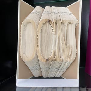 Op de afbeelding: Een boekkunst sculptuur van het woord "DONE" gemaakt door de pagina's van een boek te vouwen. De sculptuur wordt tentoongesteld in een boekenkast met andere boeken.