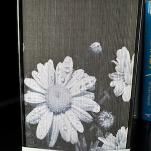 May include: A black and white book with a daisy flower design on the cover. The daisy has water droplets on the petals.