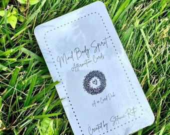 May include: A gray card deck with a white border and black text that reads "Mind Body Spirit Affirmation Cards" and "A 44 Card Deck" with a small heart graphic. The text "Created by: Stefanie Ruth" is also printed on the card.