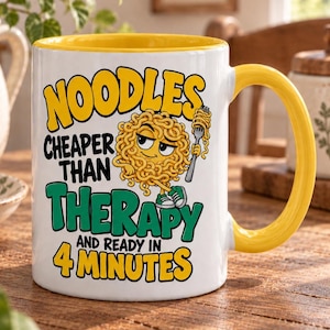 May include: A white ceramic mug with a yellow interior and handle. The front features a cartoon noodle character holding a fork, with the text: NOODLES CHEAPER THAN THERAPY AND READY IN 4 MINUTES, printed in bold yellow, black, and green lettering.
