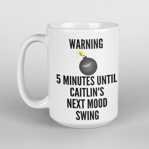 May include: White ceramic mug with a black handle. The mug features the text "WARNING" above a cartoon bomb graphic, followed by "5 MINUTES UNTIL CAITLIN'S NEXT MOOD SWING" in black text. The mug is a standard size.