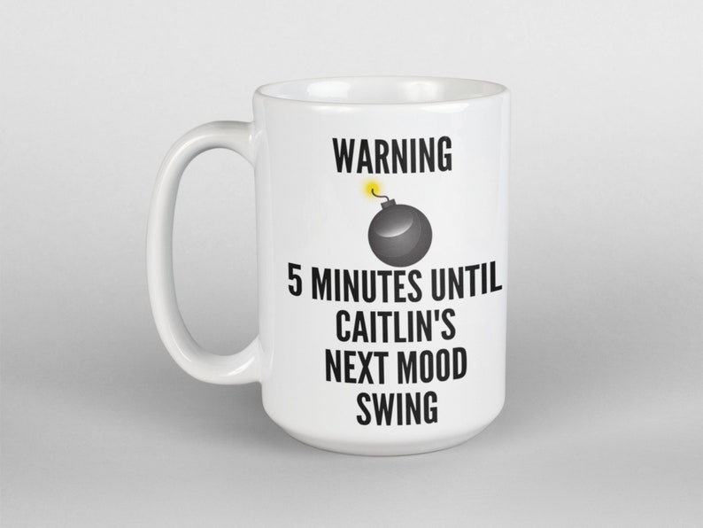 May include: White ceramic mug with a black handle. The mug features the text "WARNING" above a cartoon bomb graphic, followed by "5 MINUTES UNTIL CAITLIN'S NEXT MOOD SWING" in black text. The mug is a standard size.