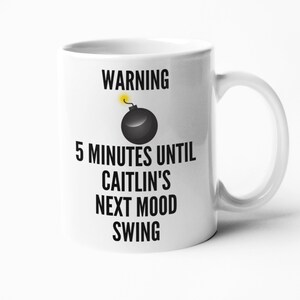 May include: White ceramic mug with a black and yellow graphic of a bomb and the text "WARNING 5 MINUTES UNTIL CAITLIN'S NEXT MOOD SWING". The mug has a curved handle.
