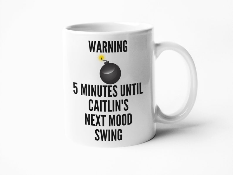May include: White ceramic mug with a black and yellow graphic of a bomb and the text "WARNING 5 MINUTES UNTIL CAITLIN'S NEXT MOOD SWING". The mug has a curved handle.