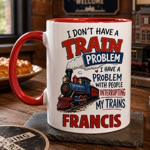 May include: A white ceramic mug with a red handle and interior rim. The mug features a graphic of a blue and red steam locomotive with smoke and the text: I DON'T HAVE A TRAIN PROBLEM I HAVE A PROBLEM WITH PEOPLE INTERRUPTING MY TRAINS FRANCIS.